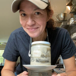 Ellie looks into the camera, smiling, over a jar of ferment