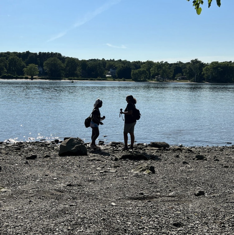 two silhouetted figures stand on the shore of a river, rough gravel and rocks on the shore and glassy wide river and blue sky behind them.