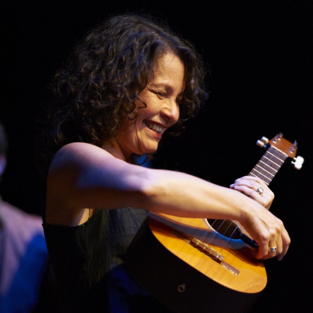 A photo of Lucía Pulido performing with an acoustic guitar. She is smiling and has the guitar on her chest.